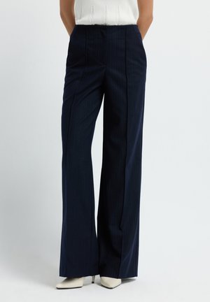 Vrouw in marineblauwe gestreepte wijde broek en witte puntige hakken voor een effen achtergrond.