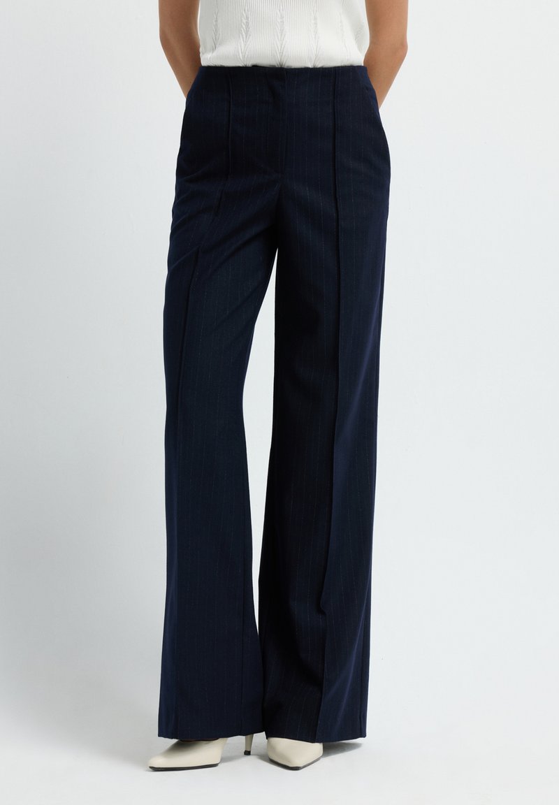 Vrouw in marineblauwe gestreepte wijde broek en witte puntige hakken voor een effen achtergrond.