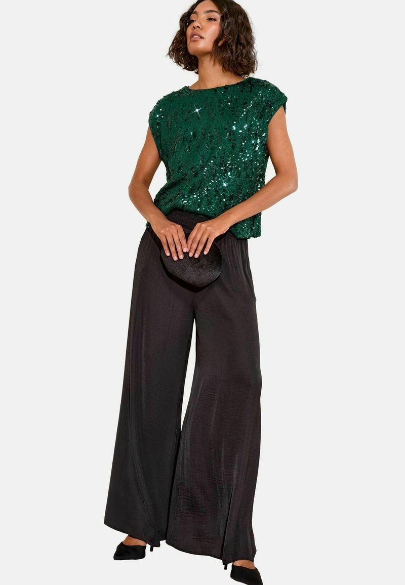 Top verde con paillettes a maniche corte abbinato a pantaloni neri larghi. La modella tiene una piccola pochette nera. Scarpe nere piatte completano l'outfit.