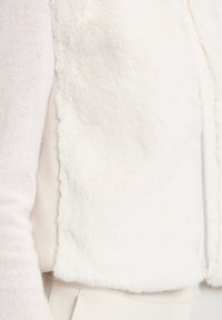 Kamizelka z sztucznego futra w kolorze kremowym o miękkiej teksturze, z zamkiem błyskawicznym i krojem do pasa, noszona na lekkim swetrze.