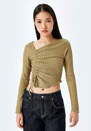 Olijfgroene cropped top met ribbelstructuur, heeft een asymmetrische halslijn en een zijband. Gecombineerd met high-waisted donkere jeans.
