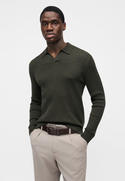 Maglione verde scuro a costine con scollo a V e colletto, abbinato a pantaloni beige chiaro e cintura marrone. Design semplice e strutturato.