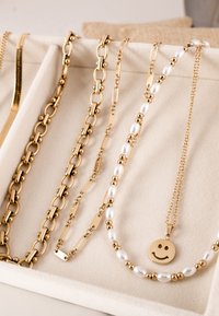 Izbor zlatnog nakita: masivne i delikatne lance, ogrlica od perlica sa zlatnim i bisernim detaljima te privjesak s licem koje se smješi.