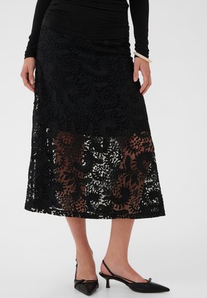 Femme portant une jupe midi noire en dentelle avec une partie inférieure transparente, un haut noir à manches longues, des escarpins noirs slingback et un bracelet blanc.