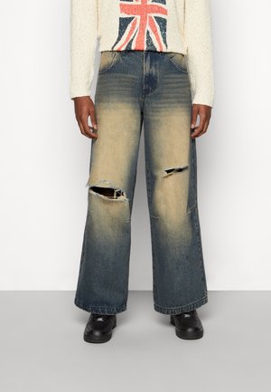 Personne portant un jean délavé à jambes larges avec de grands trous usés aux deux genoux, des chaussures noires et un pull blanc avec un motif de croix rouge et bleue.