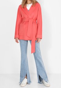 Abrigo envolvente color coral con cintura ceñida, que incluye dos bolsillos delanteros y un cuello. Combinado con vaqueros acampanados azul claro y zapatillas blancas.