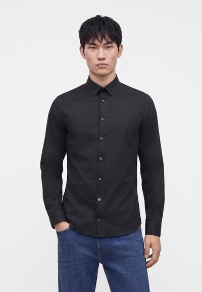 Mann trägt ein eng anliegendes schwarzes Hemd mit langen Ärmeln und blauen Jeans, steht mit einer Hand in der Hosentasche vor einem schlichten weißen Hintergrund.
