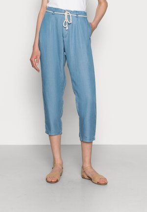 Hellblaue, knöchellange Hose mit weißem Kordelgürtel, kombiniert mit beigen, geflochtenen Sandalen mit offenem Zehenbereich an einer Person, die vor einem schlichten Hintergrund steht.