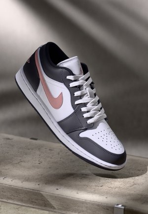 Hvid, grå og pink Nike-sneaker med snørebånd, svævende over en betongulv med blødt skygget baggrund.