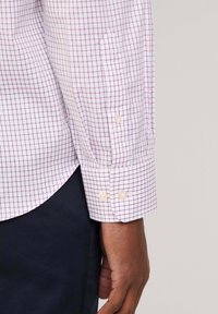 Camicia bianca con un motivo a griglia rosso e blu navy, con polsi abbottonati e un orlo leggermente curvato. Il tessuto sembra liscio e fresco.
