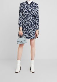 Blommig marinblå klänning med långa ärmar och krage, prydd med ljusblå blommor. Matchas med en strukturerad ljusblå handväska och vita ankelstövlar.