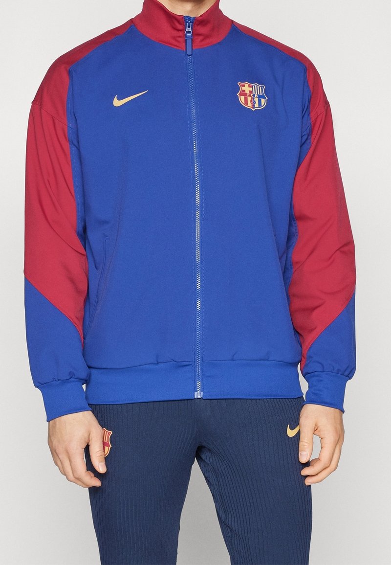 Blå och röd zip-up jacka med hög krage, med Nike-logotyp och FC Barcelona-emblem. Tillverkad av slät tyg med ribbade manschetter.