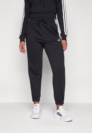 Persoon draagt zwarte Adidas trainingsbroek met elastische enkelsluitingen en witte Adidas sneakers, staand tegen een effen lichte achtergrond.