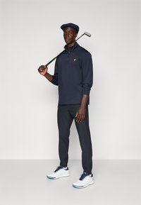 Marineblaues Langarmpolo mit Viertelreißverschluss und gelbem Logo, kombiniert mit dunklen Hosen und weißen Golfschuhen, während ein Golfschläger über der Schulter gehalten wird.