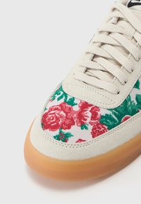 Tenisky s kvetinovou potlačou, na ktorých sú červené ruže a zelené listy na bielom plátne, doplnené béžovým semišovým špicom a podošvou z gumy.