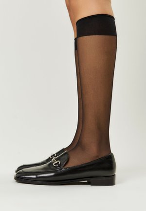 Ciorapi negri opaci, până la genunchi, asociați cu mocasini negri lucioși prevăzuți cu detalii argintii. Textură netedă și design elegant.