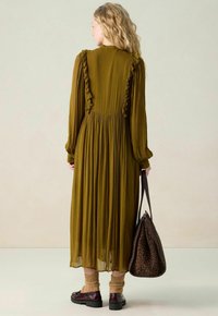 Robe longue vert olive avec des épaules volantes et une jupe plissée. Associée à des chaussettes marron et des mocassins bordeaux, tenant un sac à motif léopard.