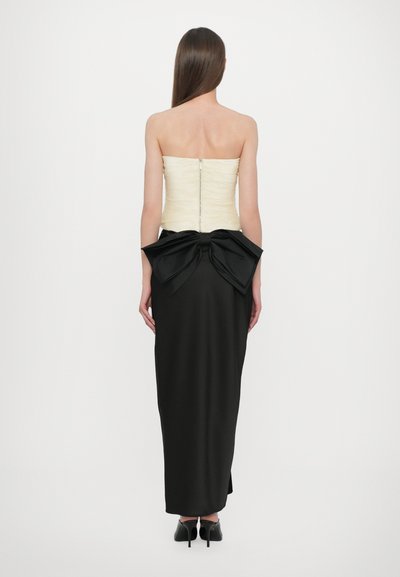 Vrouw met lang steil haar, gekleed in een strapless crème korset en een zwarte rok tot op de vloer met een grote strik aan de onderrug, staand tegen een witte achtergrond.