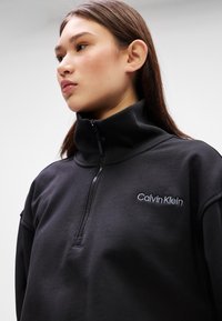 Black zip-up sweatshirt with high collar, featuring embroidered "Calvin Klein" logo in white. Smooth, soft texture and structured design.
