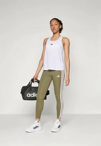 Bela atletska majica na bretele z logotipom Adidas v črni barvi, olivno zeleni pajkice z belimi črtami, črna športna torba in bele tene.