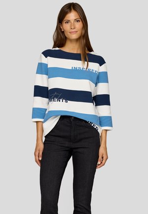 Hellblau-weiß gestreiftes Oberteil mit Dreiviertelärmeln, das die aufgestickten Wörter "INSPIRIERT" und "MOMENTE" zeigt. Kombiniert mit dunklen Jeans.