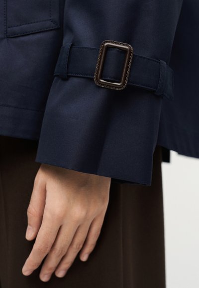 Κοντινό πλάνο ενός χεριού που ακουμπάει δίπλα σε μανσέτα ναυτικού μπλε με σκουρόχρωμο ορθογώνιο αγκράφα, σε καφέ φόντο ενδύματος.