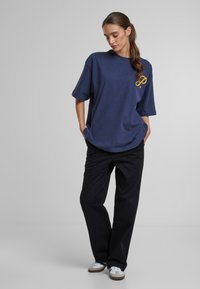 Navy oversized t-shirt with a yellow logo, short sleeves, and a rounded neckline, paired with black wide-leg pants and white sneakers.