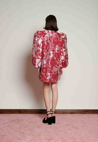 Γυναίκα που στέκεται με την πλάτη στην κάμερα, φορώντας λαμπερό ροζ φόρεμα με κόκκινα λουλουδάτα σχέδια και μαύρα ψηλοτάκουνα παπούτσια σε ροζ χαλί.