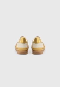 Sneakers with a cream canvas upper, gold accents, and a layered tan rubber sole. Rounded shape with a textured finish.
