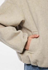 Hand rustend in de voorste zak van een beige wollen jas, gedragen met blauwe denim jeans, met details van de mouw en de tailleband.