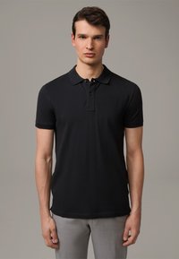 Schwarzes kurzärmeliges Poloshirt mit klassischem Kragen, Drei-Knopf-Leiste und glatter Textur. Kombiniert mit hellgrauen Hosen.
