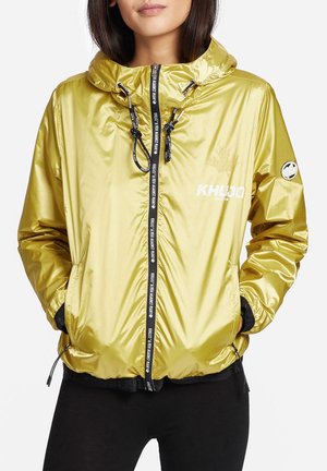 Femme portant une veste à capuche jaune brillante avec une fermeture éclair noire et des cordons, les mains dans les poches, debout devant un fond blanc.