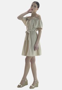Vestido beige de un solo hombro con escote con volantes, cintura ajustada y falda suelta. Combinado con sandalias de cuña doradas y una textura suave.