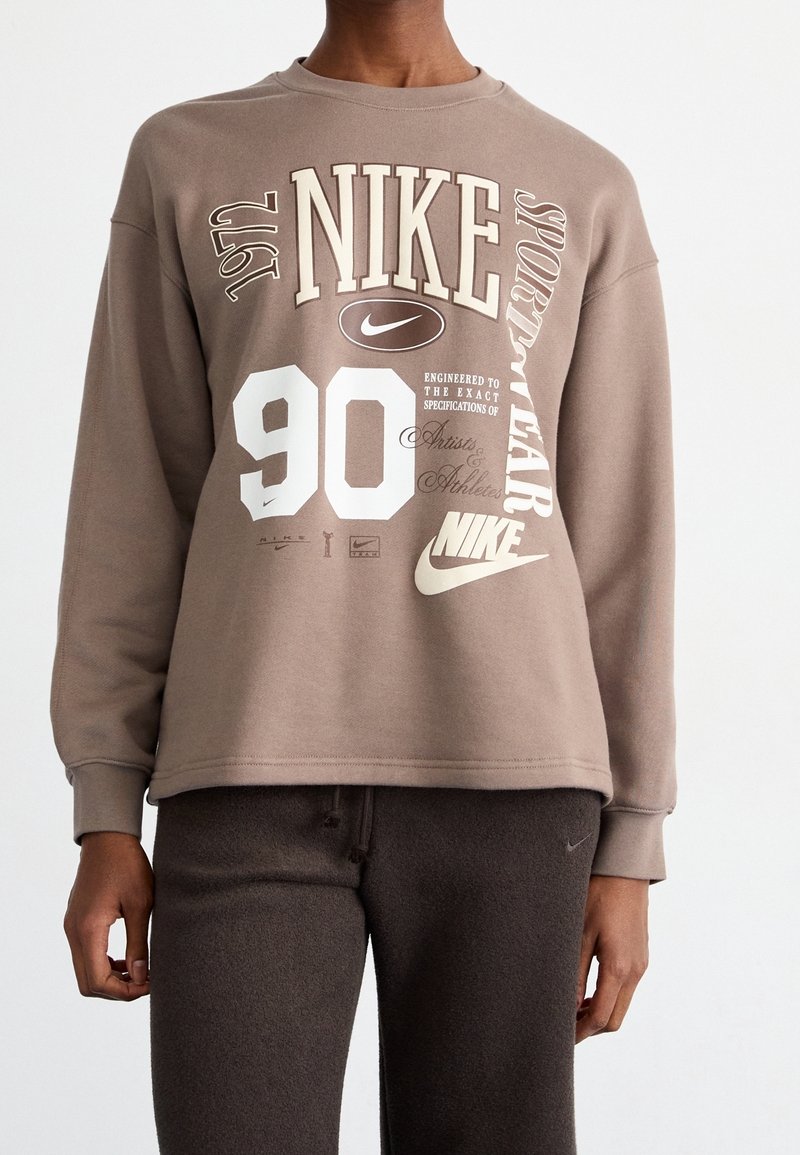 Personne portant un sweat-shirt Nike marron avec des logos et du texte blancs, associé à un pantalon marron foncé sur un fond uni.