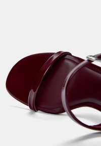 Burgundské lakované kožené sandály s širokým páskem přes přední část a štíhlým páskem na kotníku, který má malou kovovou sponu. Hladký povrch.