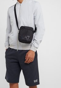 Borsa a tracolla nera in nylon con chiusura a zip, caratterizzata da un logo riflettente EA7. Forma quadrata compatta e tracolla regolabile.