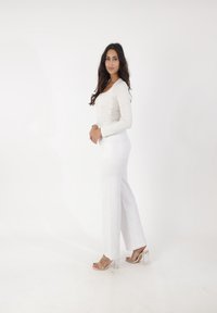 Een crèmekleurige tops met lange mouwen gecombineerd met hoge witte wijde jeans en doorzichtige gestapelde haksandalen, gepresenteerd tegen een effen witte achtergrond.