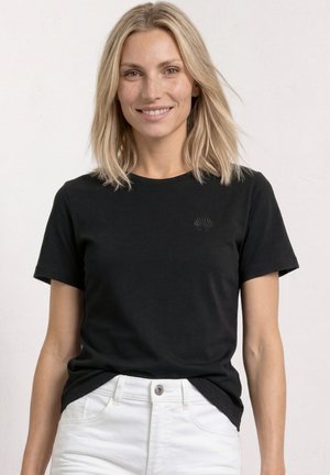 Blond kvinde iført en sort kortærmet T-shirt og hvide bukser, smilende med løst hår mod en ensfarvet lys baggrund.