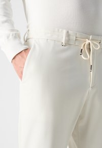 White trousers with a soft texture, featuring a drawstring waist and subtle pockets, shown with a hand inserted in the pocket.
