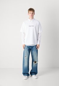 Balta trumpa rankovėmis marškiniai su "Calvin Klein" logotipu, dėvimi ant baltų ilgomis rankovėmis marškinių; nudėvėti šviesiai mėlyni plataus kirpimo džinsai; balti sportbačiai.