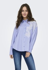 Camisa azul riscada com detalhes em renda em um dos ombros, mangas longas e um corte descontraído. Usada com jeans azul escuro.