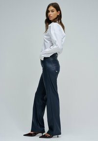 Pantalon large en simili cuir bleu foncé avec des détails de couture visibles, associé à une chemise blanche à boutons et des talons aiguilles.