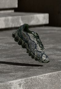 Teniska s tmavě zeleným a béžovým hadím vzorem, černou bublinovou podrážkou a tmavě zelenými tkaničkami, vznášející se nad betonovým povrchem s pozadím schodů.