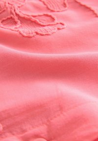 Tejido de color coral-pálido con una textura suave, que presenta intrincados patrones florales bordados y sutiles detalles de costura.