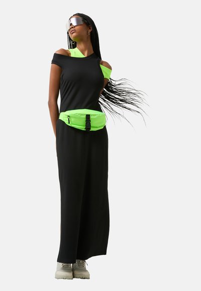Zwarte off-shoulder jurk met ribbelstructuur, gecombineerd met een neon groene heuptas en grijze sneakers. Lang haar waait in de bries.
