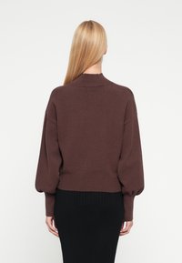 Brązowy dziergany sweter z wysokim kołnierzem, opuszczonymi ramionami i ściągaczem u dołu. Tylny widok ukazuje detale mankietów w formie balonów.