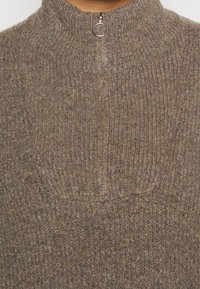 Pull en maille dans des tons marron atténués, avec un col montant et une fermeture éclair quart avec un anneau métallique. Motif côtelé texturé sur l'ensemble.