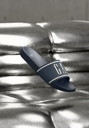 Sandale à glissière bleu marine avec logo GAP blanc sur la sangle, flottant au-dessus d'une surface capitonnée argentée métallisée brillante.
