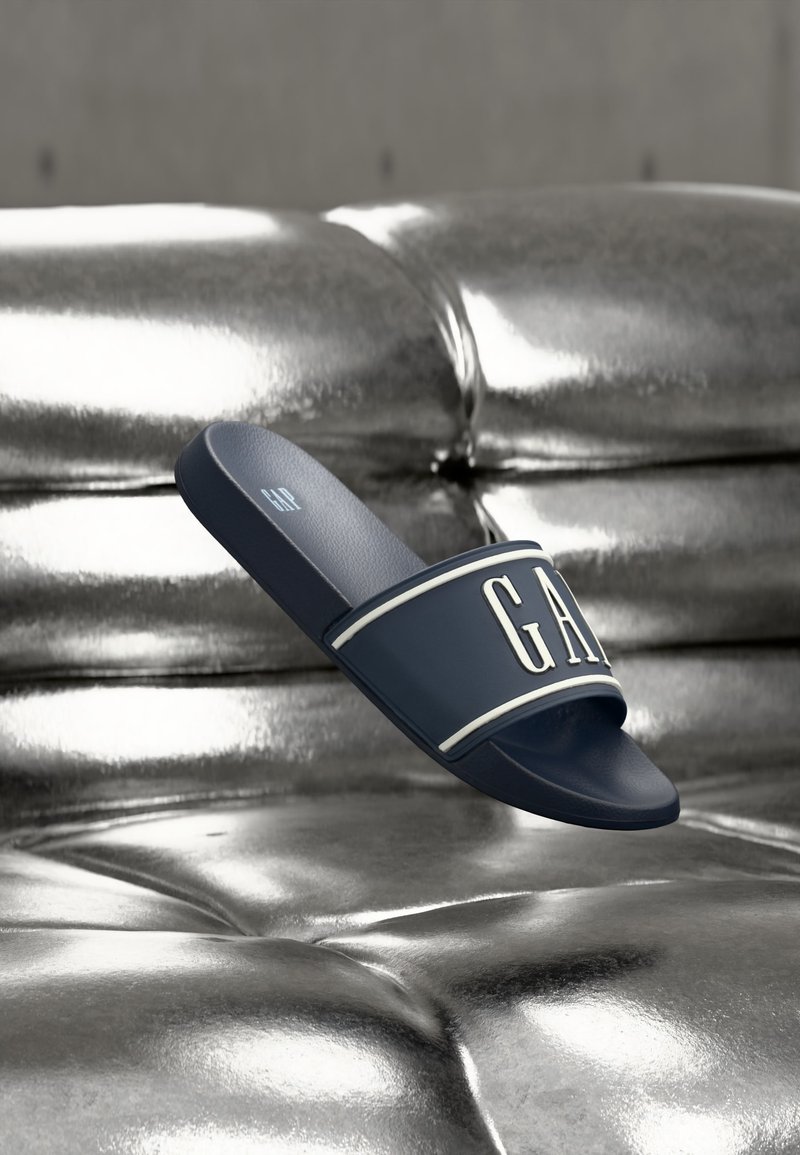 Sandale à glissière bleu marine avec logo GAP blanc sur la sangle, flottant au-dessus d'une surface capitonnée argentée métallisée brillante.