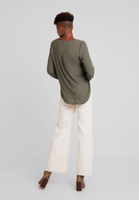 Olivegrünes Langarmhemd mit Knopfdetails am Rücken, kombiniert mit cremefarbenen weitem Hosen und gemusterten Schuhen.